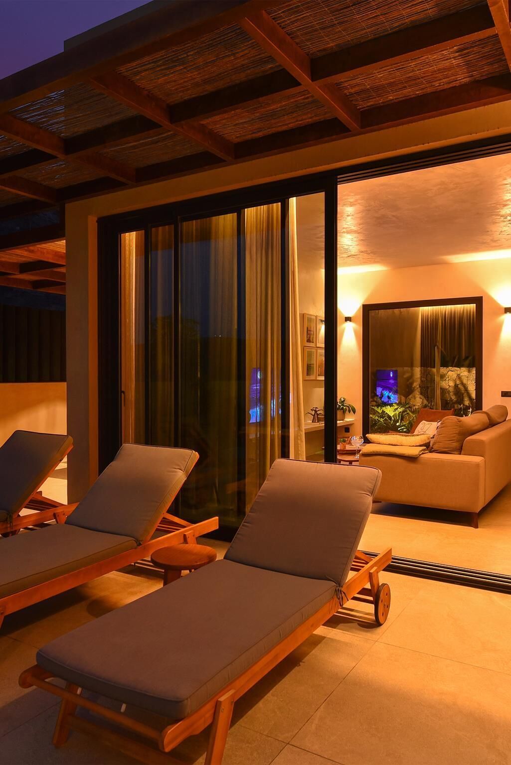 A modern outdoor lounge area with two sun loungers and a sofa, a glass door leading to a living space, and a thatched roof structure.