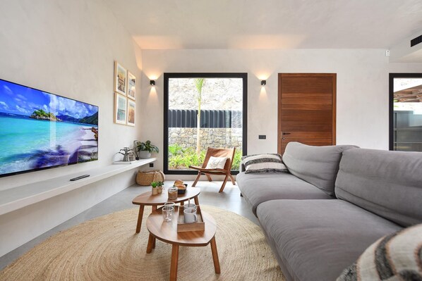 A modern living room with a flat-screen TV, a grey sofa, a wooden coffee table, and a view of a stone wall and greenery through a glass door.