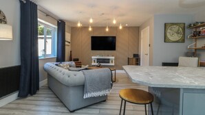 A modern living room with a grey sofa, a wooden accent wall, a flat-screen TV, and a kitchen area with a marble countertop. - Ty Glan Morfa (Porthmadog)
