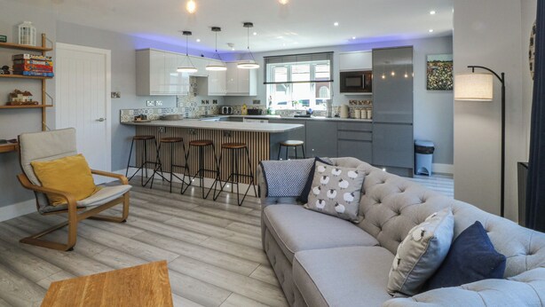 A modern kitchen with bar stools, a sofa with patterned cushions, and pendant lighting. - Ty Glan Morfa (Porthmadog)