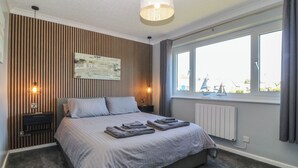 A bedroom with a bed, two bedside tables, a wall-mounted lamp, a window with a view, and a radiator. - Ty Glan Morfa (Porthmadog)
