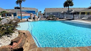 A clear blue pool surrounded by a white fence, palm trees, and buildings with a thatched roof. - `Accessible and Pet-Friendly: 2 Community Pools and a 1-Level Flat Entrance for Ultimate Comfort` (Port Aransas)