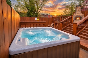 A hot tub with a stone surround, wooden deck, and a stone fireplace.