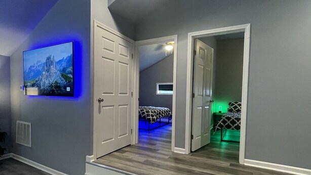Un pasillo moderno con un televisor de pantalla plana montado en la pared, suelo de madera y una escalera que conduce a un dormitorio con una cama e iluminación ambiental verde.