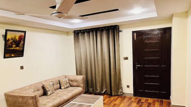 A living room with a sofa, a coffee table, a ceiling fan, a door, and a painting on the wall. - Comfy apartment in Bahria town civic center. (Rawalpindi)
