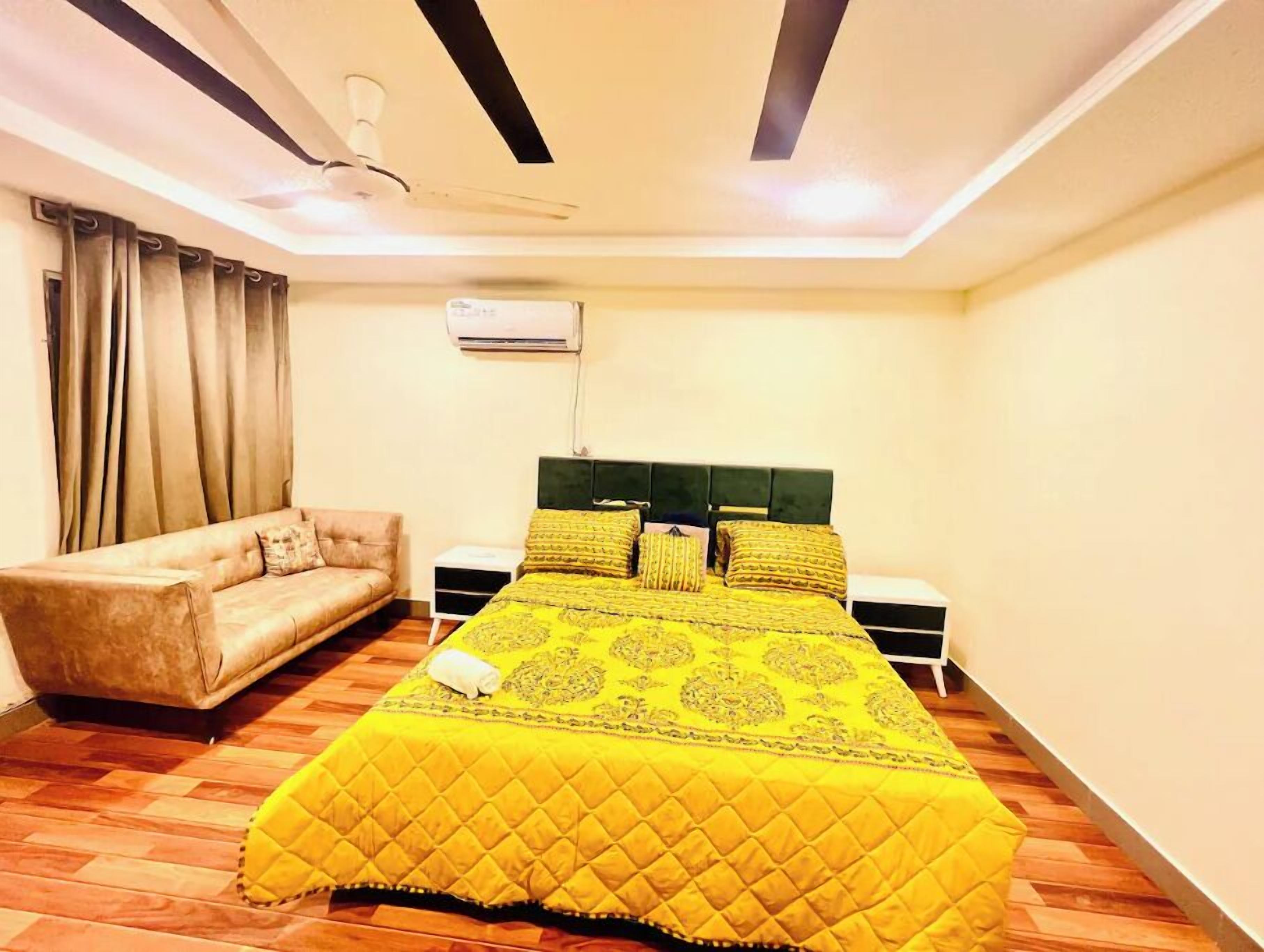 A bedroom with a bed, a sofa, a nightstand, an air conditioning unit, and a ceiling fan.