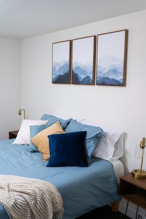 A neatly made bed with blue and white bedding, a wooden nightstand with a lamp, and three framed pictures on the wall.