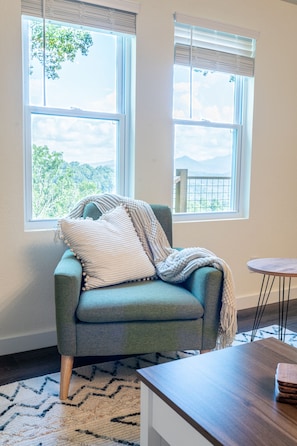 A teal armchair with a white and beige throw blanket, positioned in front of a window with a view of trees and mountains.