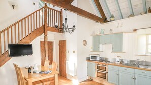 A spacious kitchen with wooden flooring, a dining area with a wooden table and chairs, a staircase with wooden railings, and a large window providing natural light. - Dovecote (St Austell)