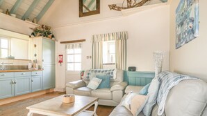A cozy living room with a sofa, a coffee table, and a kitchen area with cabinets and a window. - Dovecote (St Austell)