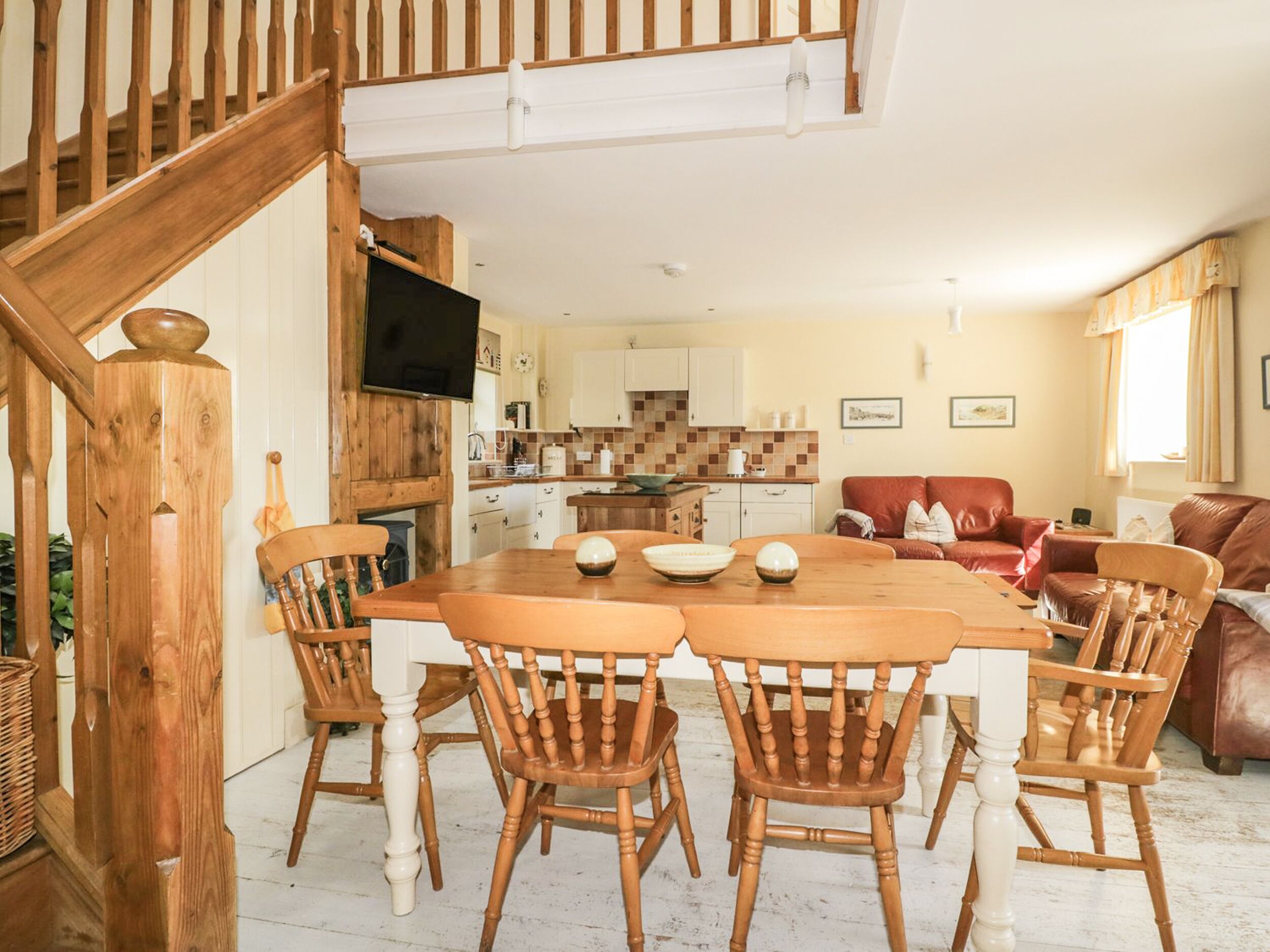 A dining area with a wooden table and chairs, a kitchen in the background, and a staircase leading to an upper level.