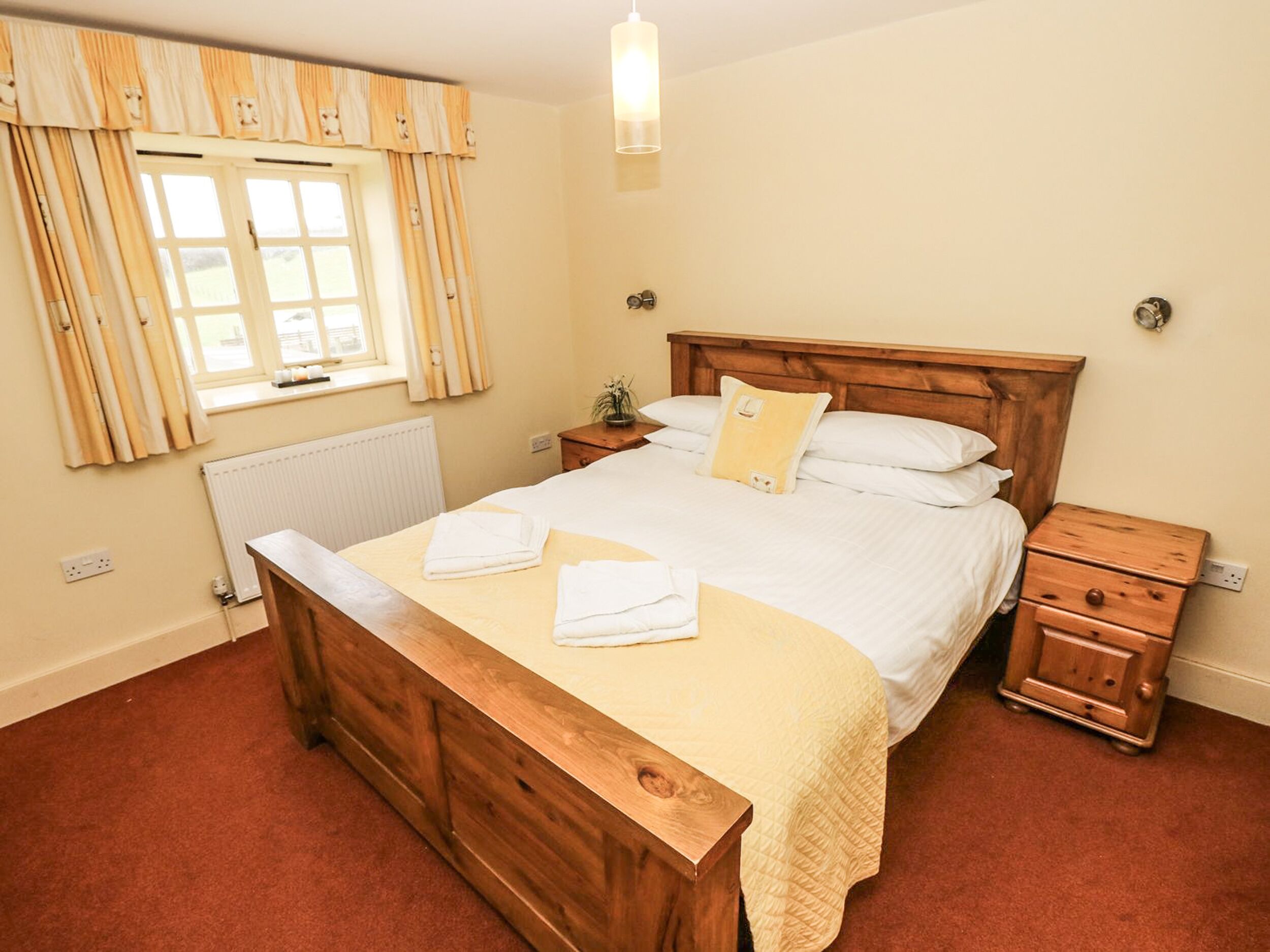 A bedroom with a wooden bed, a bedside table, a nightstand, a window with curtains, and a radiator.