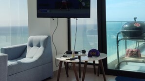 A modern living room with a large flat-screen TV, a comfortable sofa, a small round table, and a view of the ocean through a large window. - Ocean Front Suite Apartment in Manta Poseidon Building (Manta)