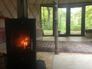 Une pièce chaleureuse avec un poêle à bois, un lit recouvert d’une courtepointe à motifs floraux et une vue sur une forêt, visible à travers de grandes portes vitrées.