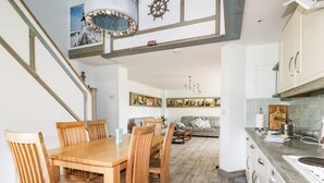 Un coin repas avec une table et des chaises en bois, une cuisine avec des meubles blancs et un escalier agrémenté d’un plafond décoratif.