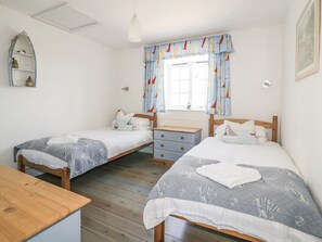 A bedroom with two single beds, a wooden dresser, a small table, and a window with curtains. - Owls Roost (St Austell)