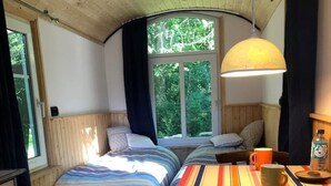 A cozy room with two beds, a table with a striped tablecloth, a lamp, and a window with a view of trees. - Construction trailer - in the middle of nature (Kleve)
