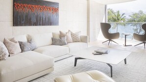 A modern living room with a white sofa, a coffee table, and two armchairs. There is a large painting on the wall and a view of the outdoors through large windows.