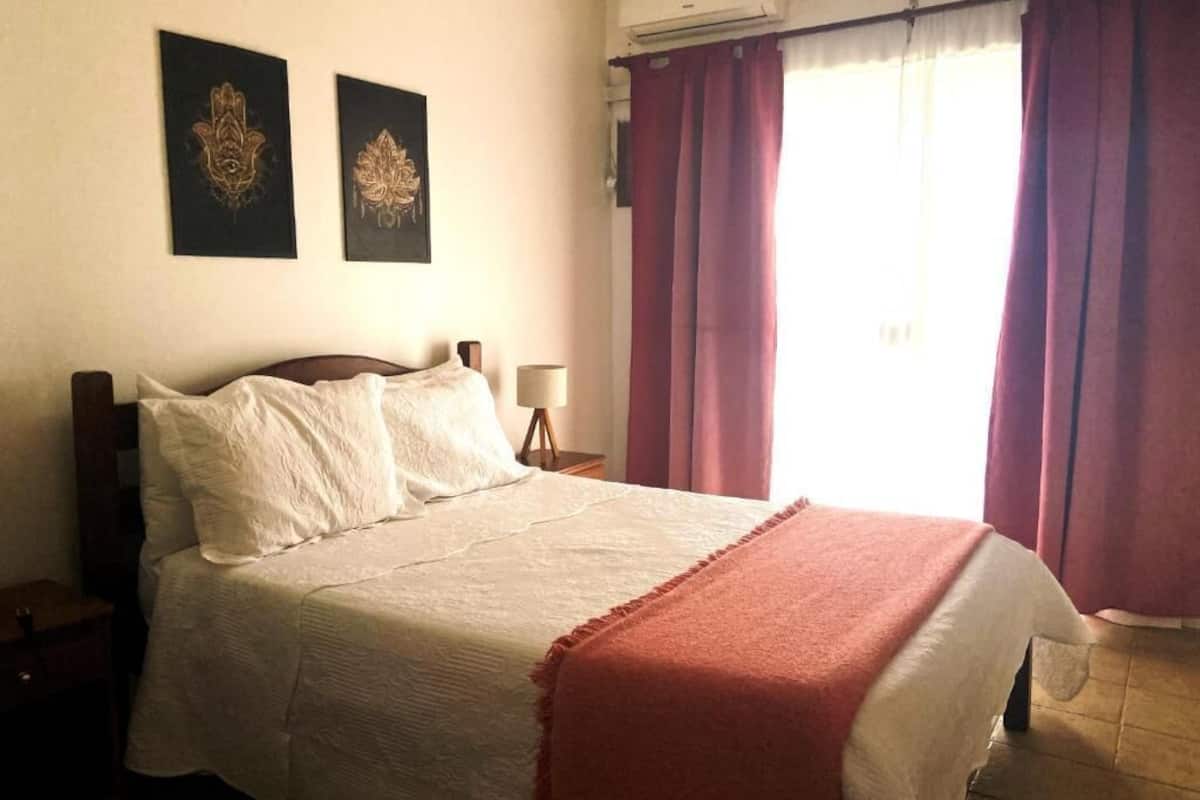 Ένα υπνοδωμάτιο με ένα κρεβάτι, ένα κομοδίνο, ένα φωτιστικό, δύο διακοσμητικά στοιχεία στον τοίχο και ένα παράθυρο με κουρτίνες.