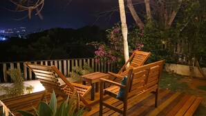 Eine Holzdielen-Terrasse mit Tisch und Stühlen, umgeben von Pflanzen und einer Palme, mit Blick auf eine nächtliche Skyline.