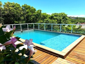 Une piscine rectangulaire dotée d’une terrasse en bois et d’une balustrade blanche, entourée d’une végétation luxuriante et surplombée par un ciel bleu clair.