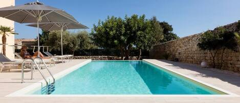 A rectangular swimming pool with clear blue water, surrounded by a stone wall and trees. There are lounge chairs and an umbrella near the pool.