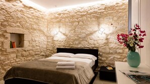 Un dormitori amb paret de pedra, un llit amb capçaleri fosca, una petita taula de nit i un vas amb flors.