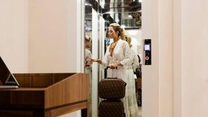 Una dona amb un vestit blanc dret a un passadís, amb una maleta i un ordinador portàtil sobre una taula.