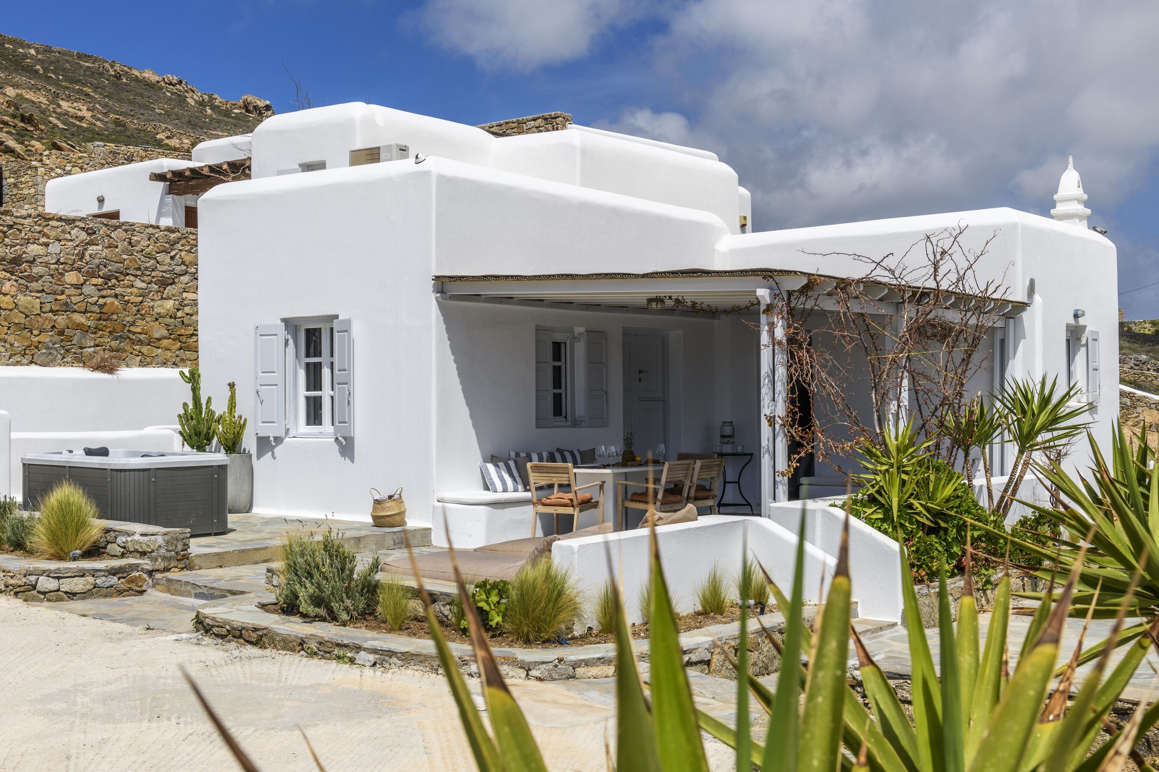 A white building with a stone wall, a hot tub, and a patio with chairs and a table.