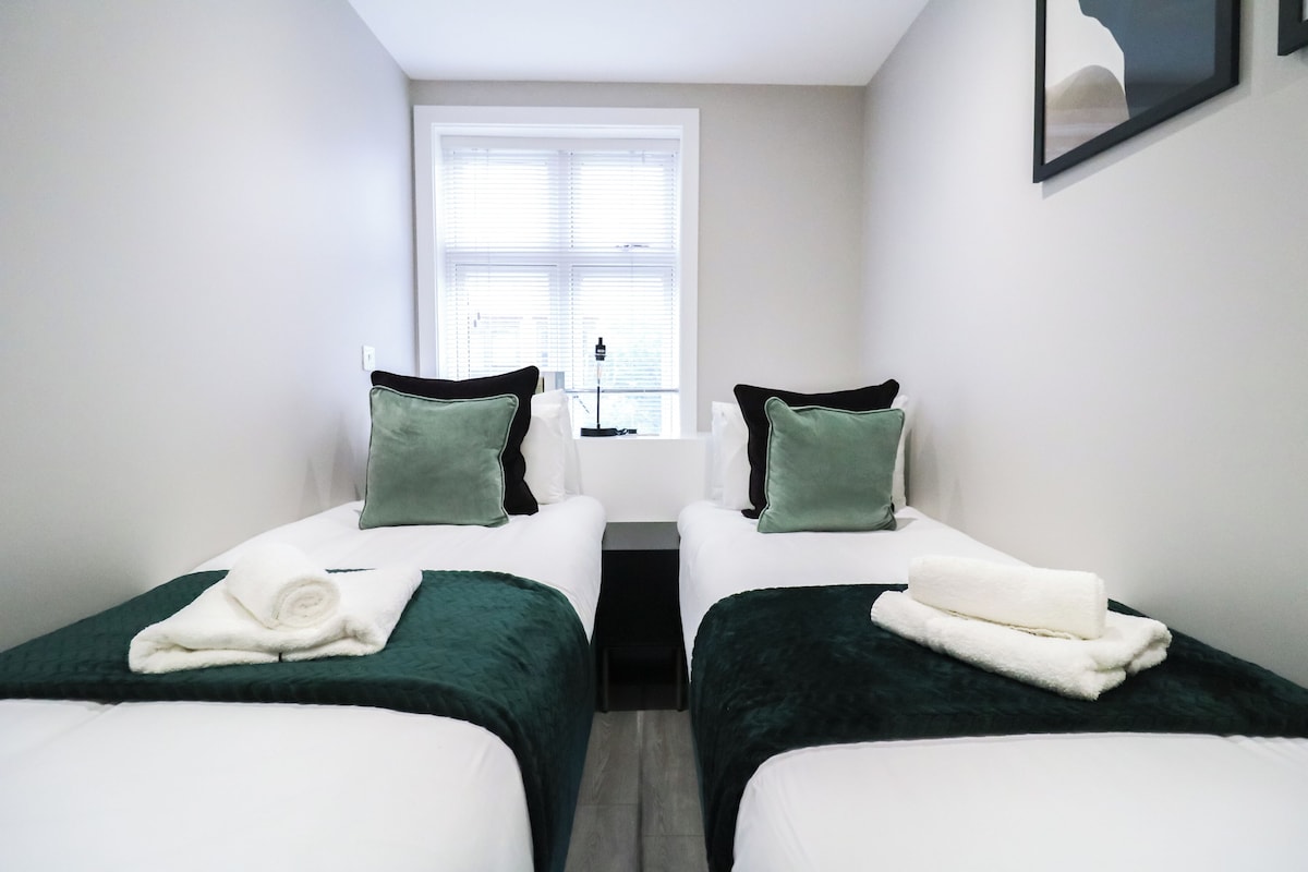 A hotel room with two single beds, each with a green pillow and a white towel.