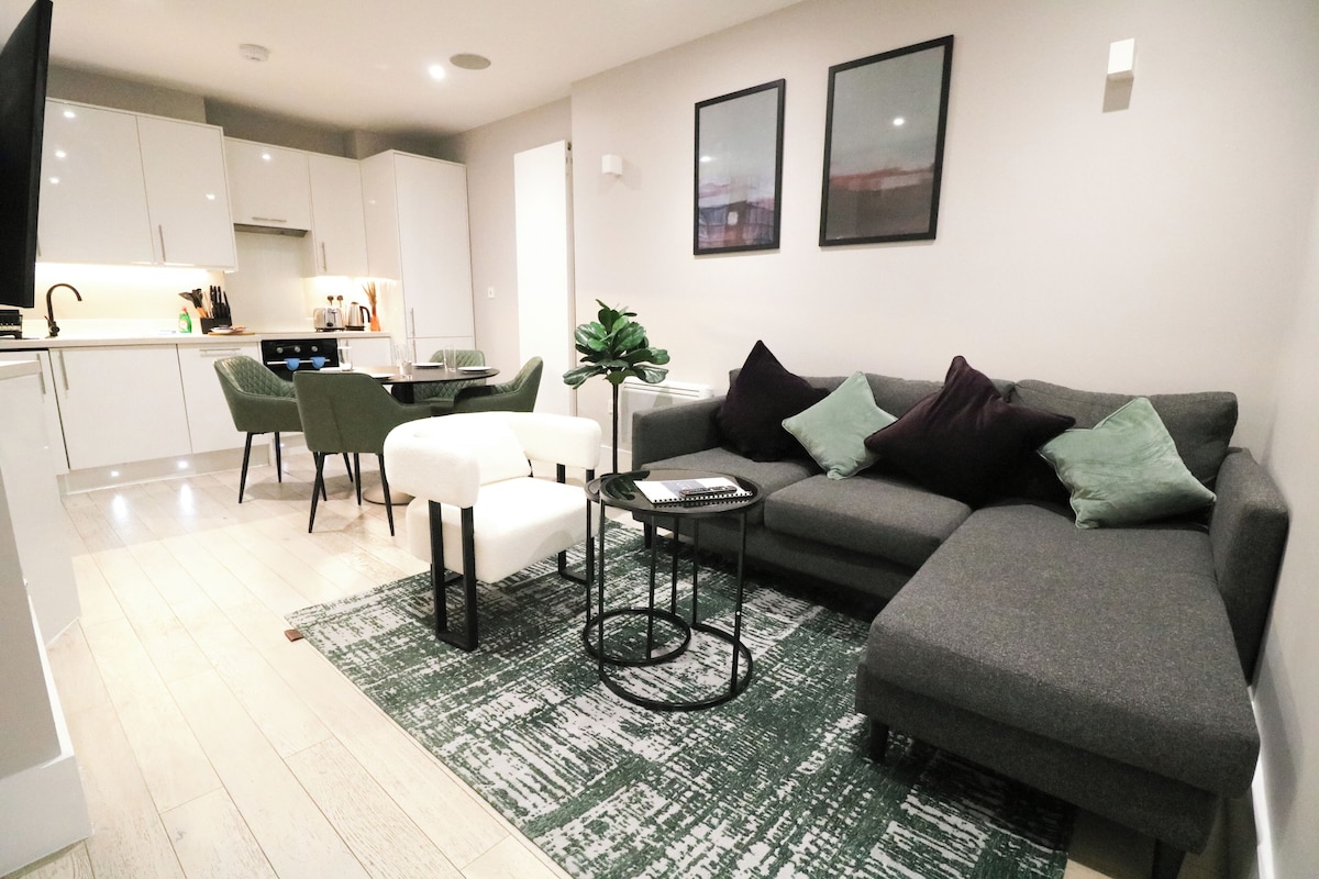 A modern living room with a grey sofa, a small side table, and a green armchair. There is a kitchen area with white cabinets and a dining table with chairs.