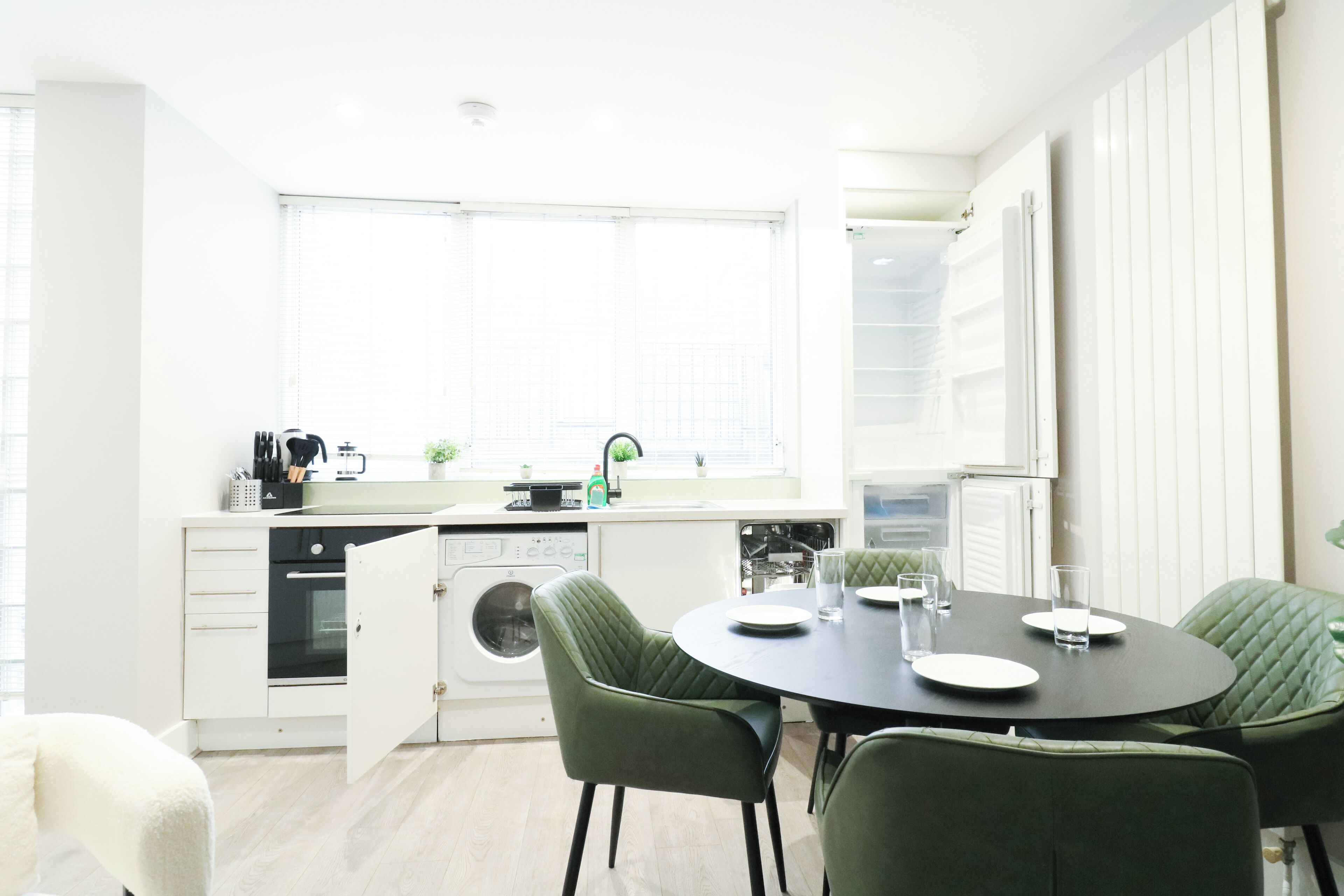A modern kitchen with a dining area, featuring a round table, green upholstered chairs, and a built-in oven.