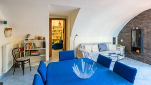 A modern living room with a blue sofa, a glass coffee table, and a bookshelf. There is a dining area with a blue table and chairs. The room has a curved ceiling and a stone wall.