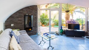 A living room with a stone wall, a glass table, a sofa, a TV, and a view of a garden through large glass doors.