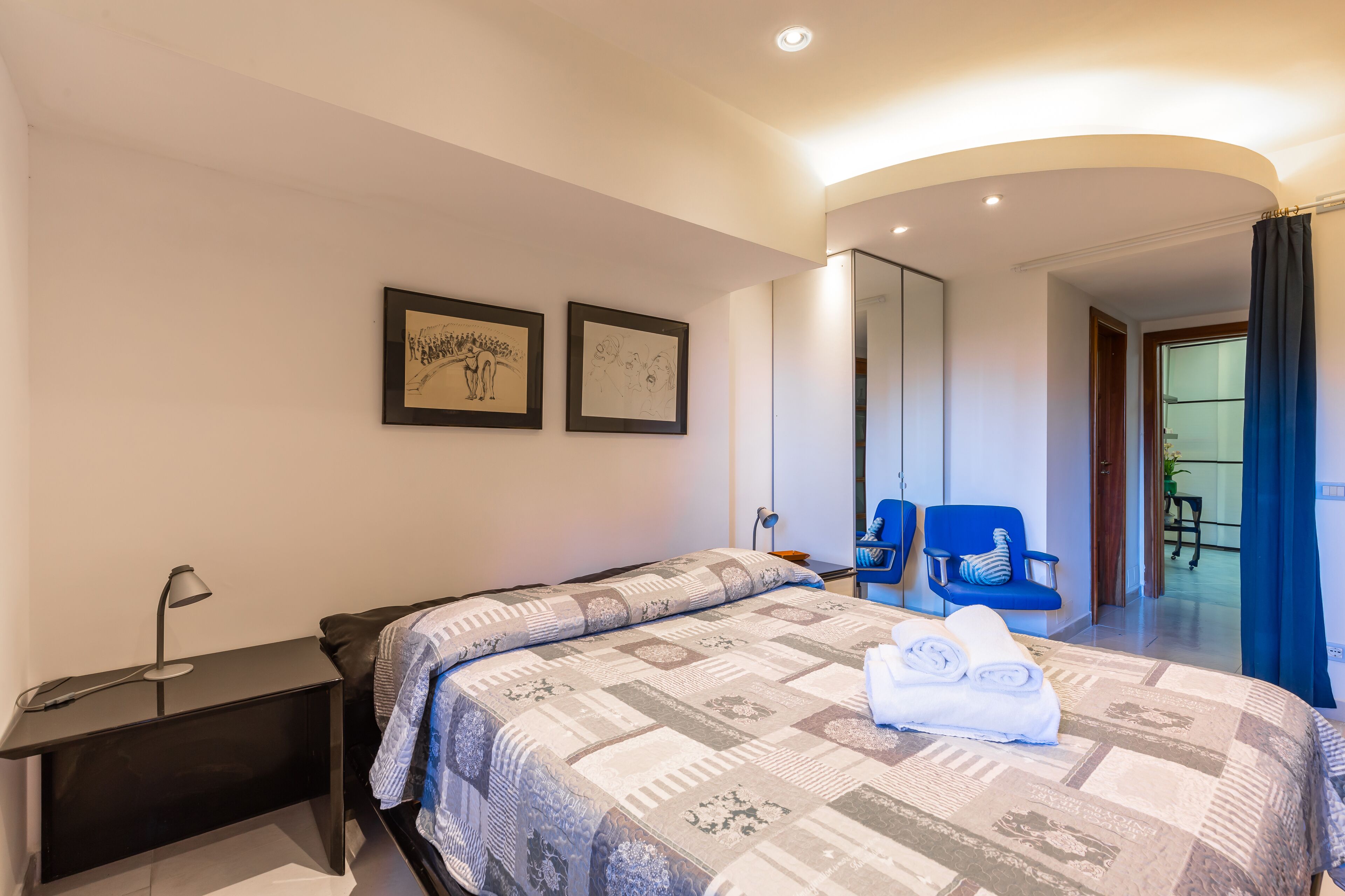 Een moderne hotelkamer met een groot bed, een nachtkastje, een bureaulamp, twee ingelijste kunstwerken aan de muur, een blauwe stoel en uitzicht op de gang en een andere kamer.
