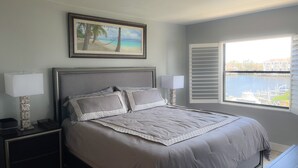 A bedroom with a large bed, two bedside lamps, a nightstand, a framed picture of a beach, and a window with a view of a marina. - Remodeled Condo, Steps to the Beach (Naples)