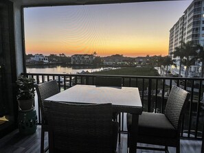 A balcony with a table and chairs overlooking a body of water and buildings during sunset.