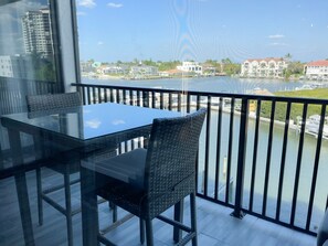 A balcony with a glass table and wicker chairs, overlooking a marina with boats and buildings.