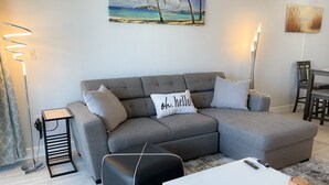 A modern living room with a grey sectional sofa, a black ottoman, a white coffee table, and a painting of a beach scene. - Remodeled Condo, Steps to the Beach (Naples)