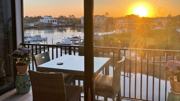 A balcony with a table and chairs, overlooking a marina with boats and buildings in the background during sunset. - Remodeled Condo, Steps to the Beach (Naples)