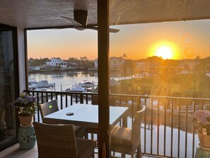 Un balcon avec une table et des chaises, donnant sur une marina où l’on aperçoit des bateaux et des bâtiments en arrière-plan, au moment du coucher du soleil.