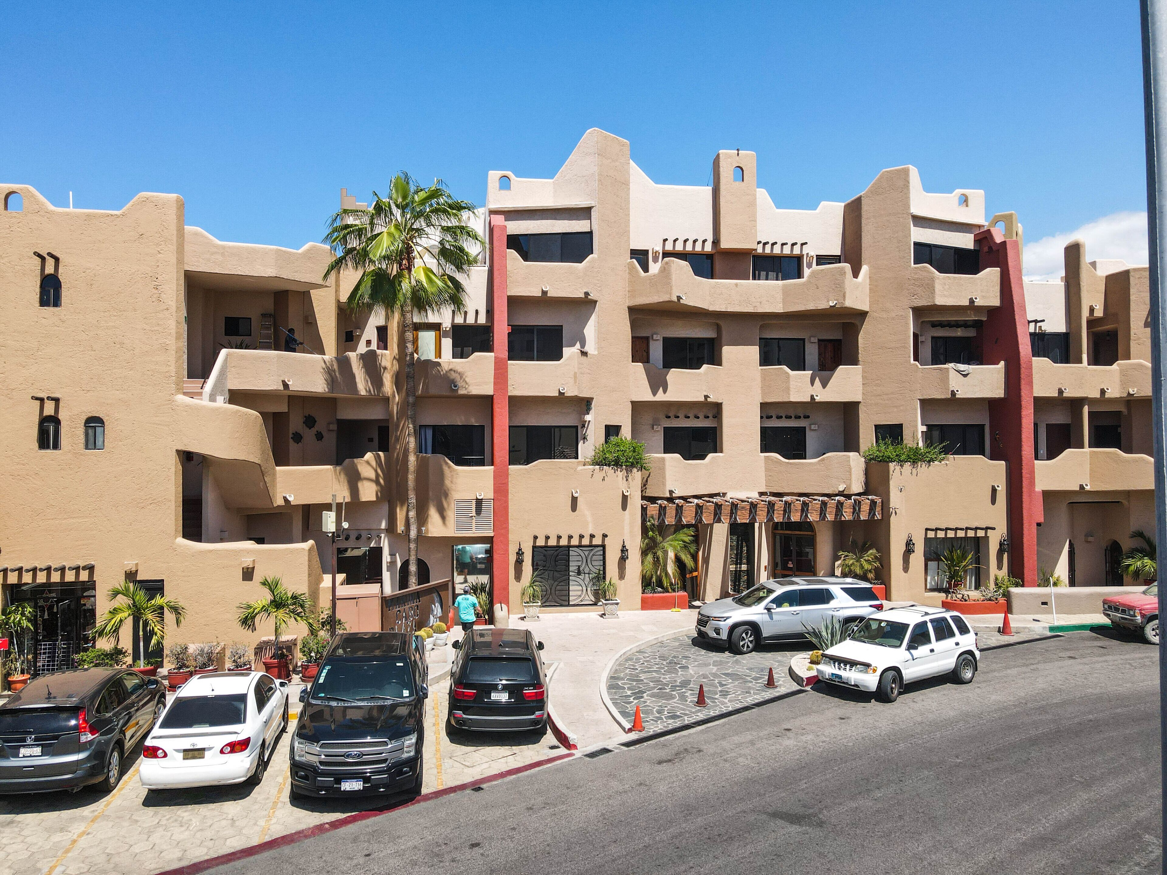 A multi-story building with a beige facade, balconies, and a parking area with several cars.