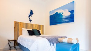 Un dormitori amb un capçal de llit de fusta, un llit amb roba de llit blanca, una banca blava i una pintura mural d'una onada.
