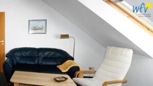 A small, cozy room with a black leather sofa, a wooden table, a white armchair, and a wooden cabinet. There is a door and a framed picture on the wall. - Haus Sleeboom - mit 53 qm in Ruhiger Lage Gelegen (Borkum)