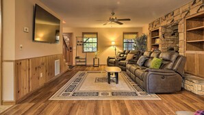A living room with a wooden floor, a sofa, a coffee table, a TV mounted on the wall, and a stone fireplace. - Spacious cabin with AC, private lake, hot tub, movie room (Arnold)