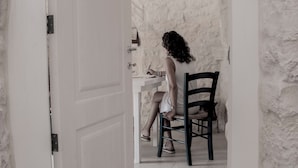 A room with a white door, a stone wall, and a person sitting at a table. - trullo manivida (Puglia)