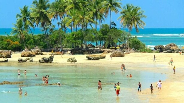 A beach with clear turquoise water, palm trees, and people enjoying the sand and water. - Pousada Camburi (Cairu)