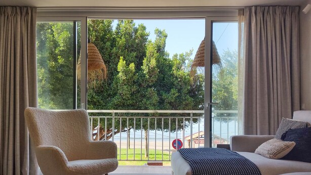 Moderni hotellihuone, jossa on suuri liukuovi, joka johtaa parvekkeelle. Huoneessa on myös sohva, nojatuoli ja pieni sohvapöytä, jonka päällä on kynttilöitä.