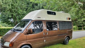 Una furgoneta camper marrón estacionada sobre césped, con un bosque al fondo.
