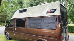 Furgoneta camper marrón estacionada sobre césped, con una dirección web a la vista.
